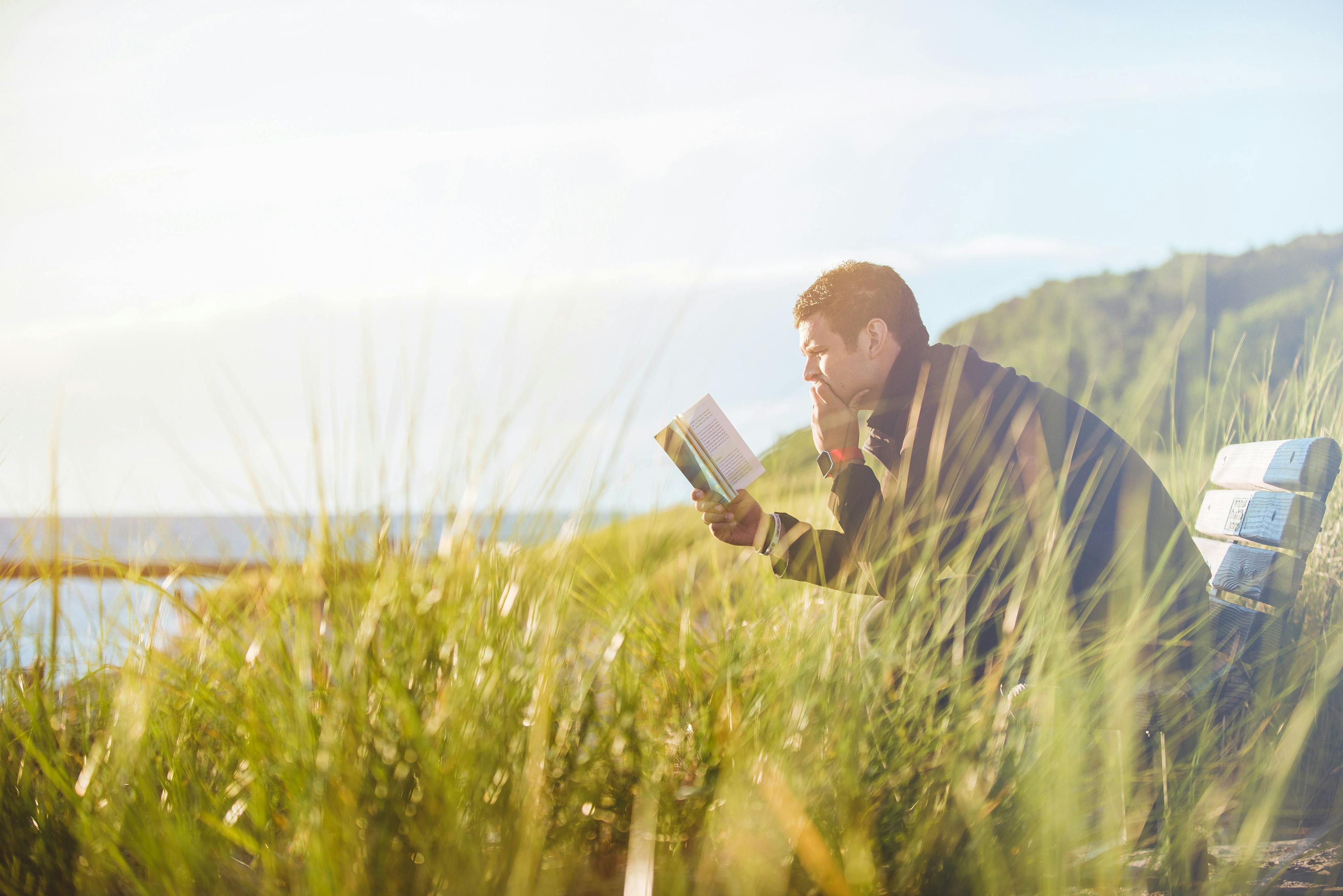 Hombre de mediana edad leyendo en la naturaleza, está sentado sobre un banco, rodeado de vegetación en un claro, frente al mar en un día soleado.