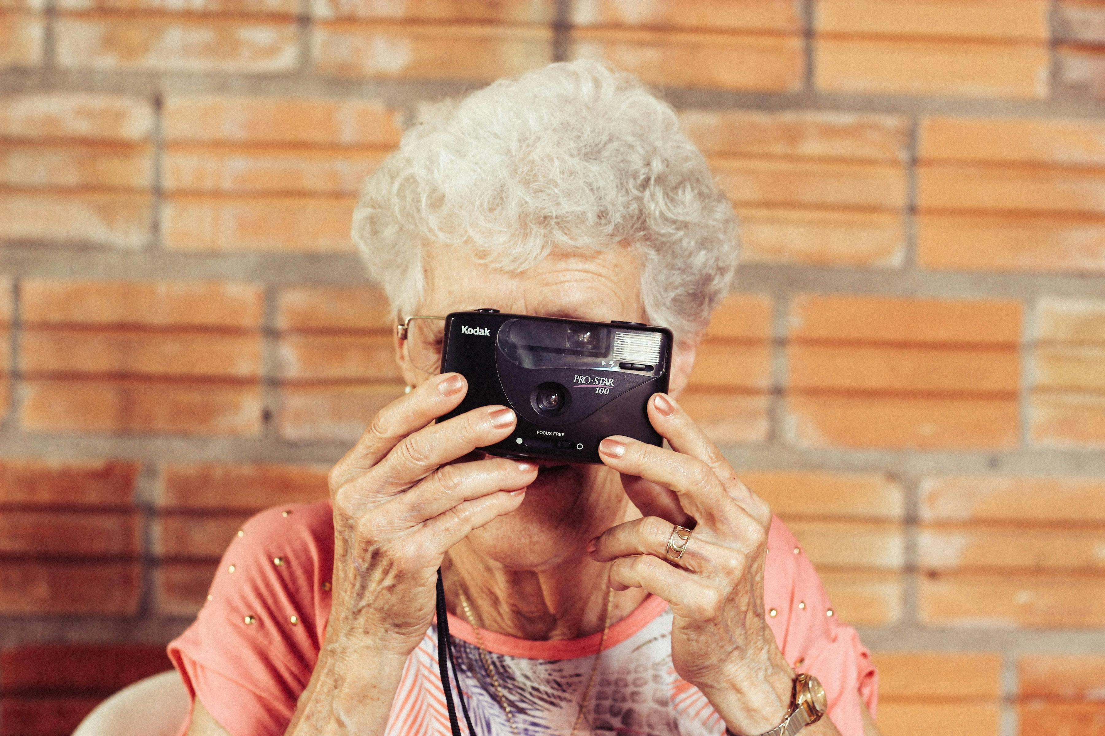 Mujer en edad madura sostiene una cámara analógica frente a su rostro, podemos ver en detalle sus arrugas, sus uñas esmaltadas y cabello blanco. La imagen tiene tonos cálidos y evoca alegría, resiliencia y nostalgia.