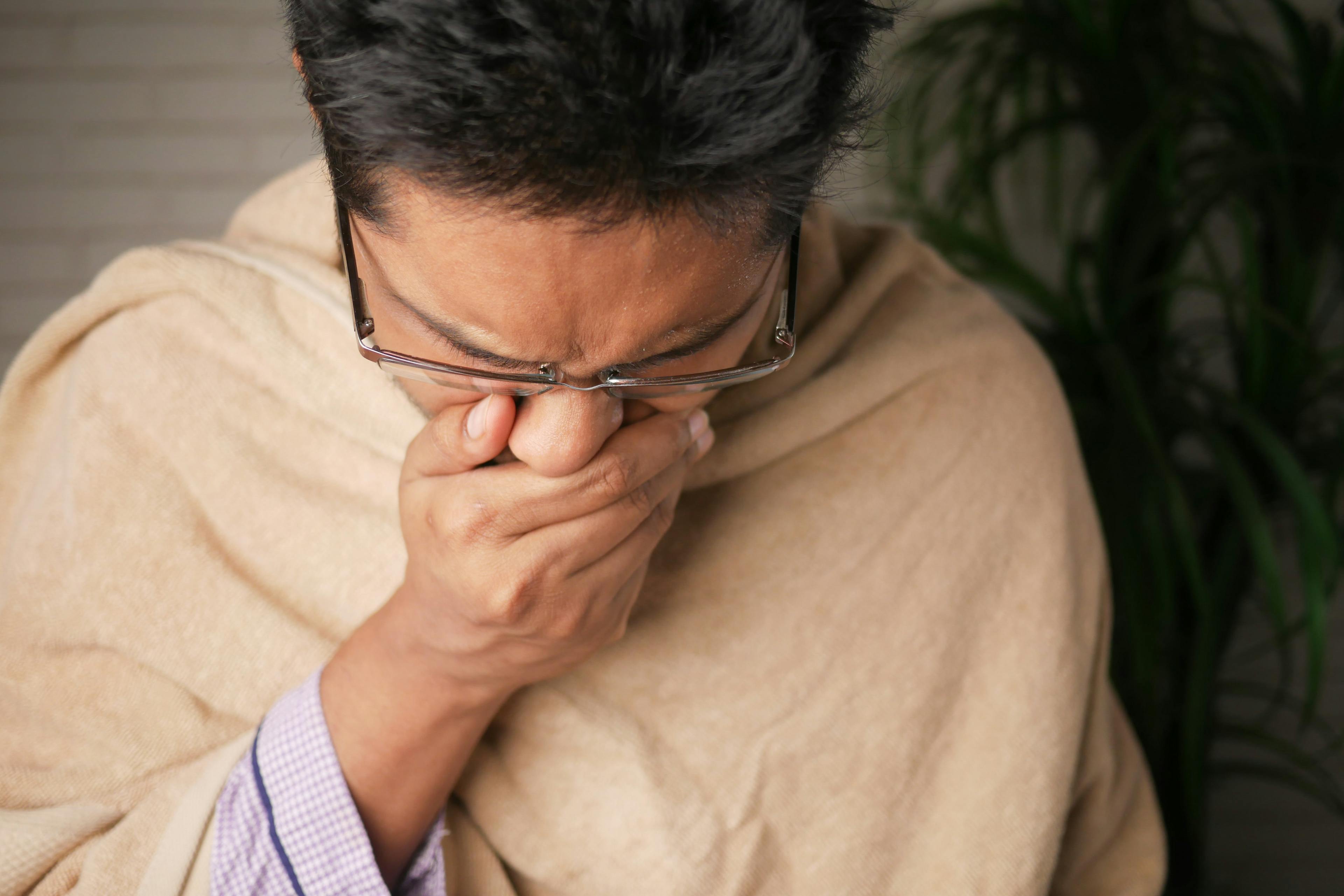 Hombre joven con rasgos asiáticos tapándose la boca para toser y con expresión de dolor