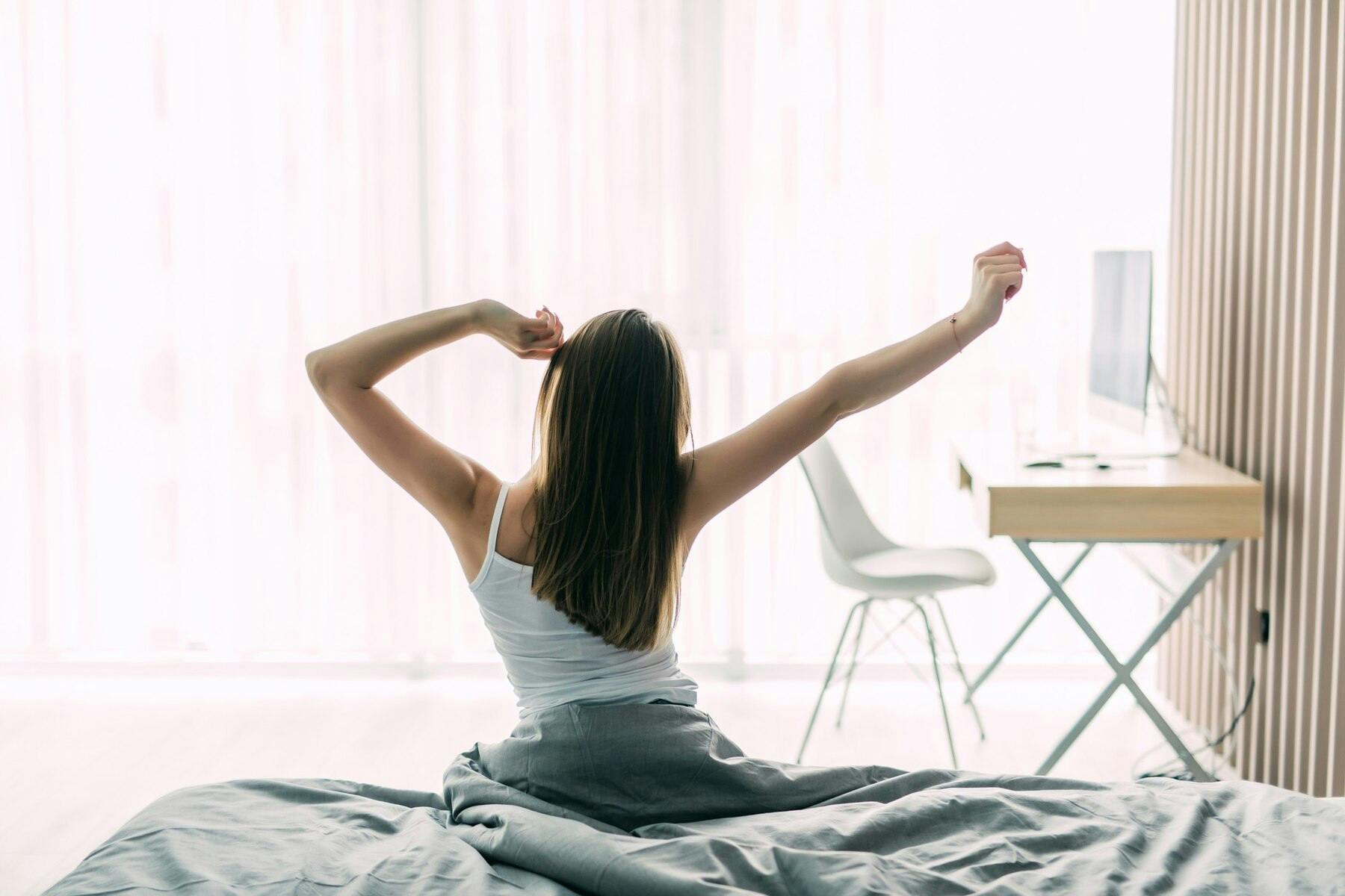Joven mujer de espaldas desperezándose luego de despertar, sentada sobre su cama, sábanas blancas, luz clara de la mañana
